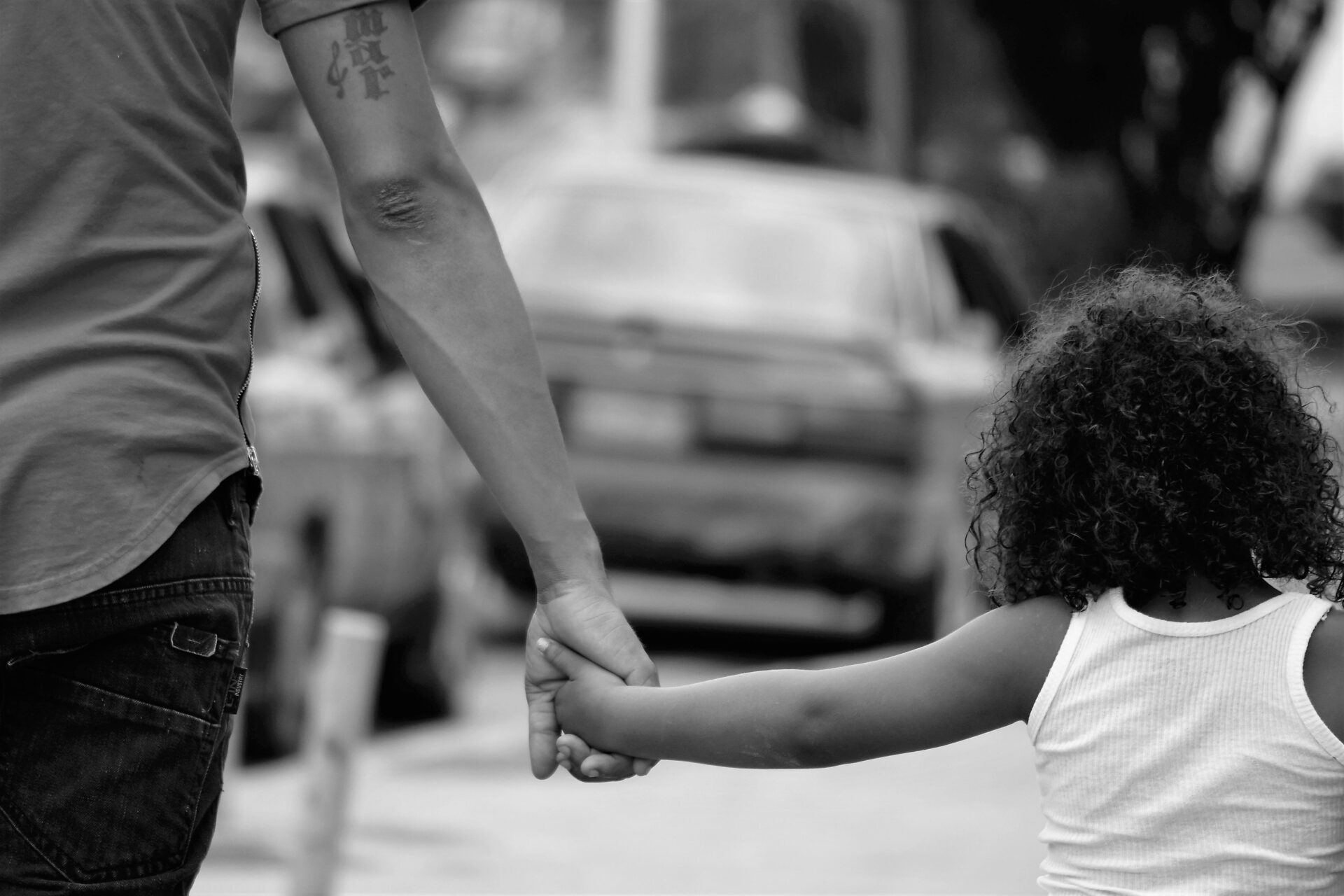 A black and white photo of a man and child holding hands on an urban street.
