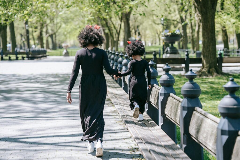 A mother and daughter in matching outfits enjoy a sunny walk in a lush green park.