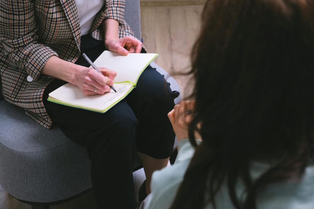 From above of crop anonymous psychotherapist taking notes in agenda while speaking with woman during psychological session