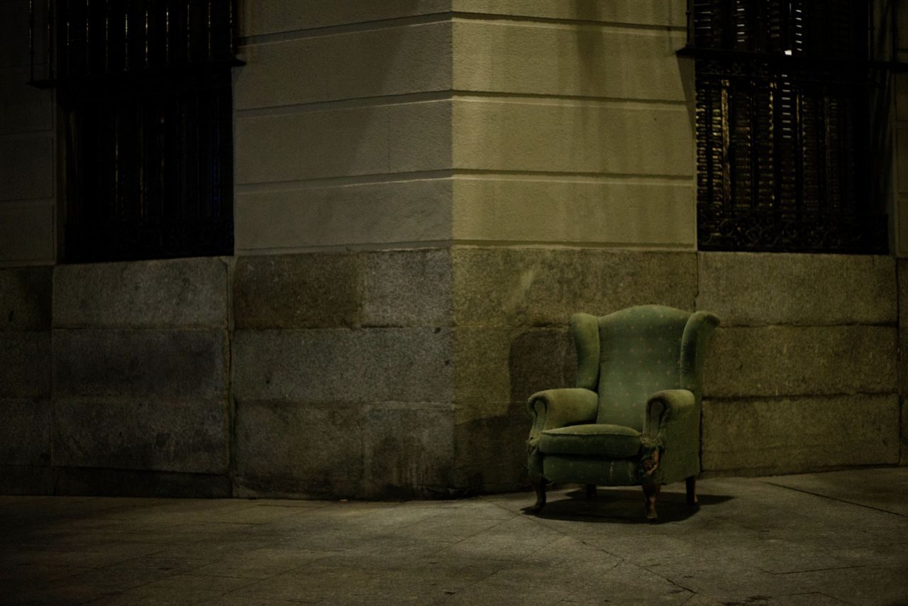 An old green armchair sits abandoned against a dark city wall at night in Madrid.