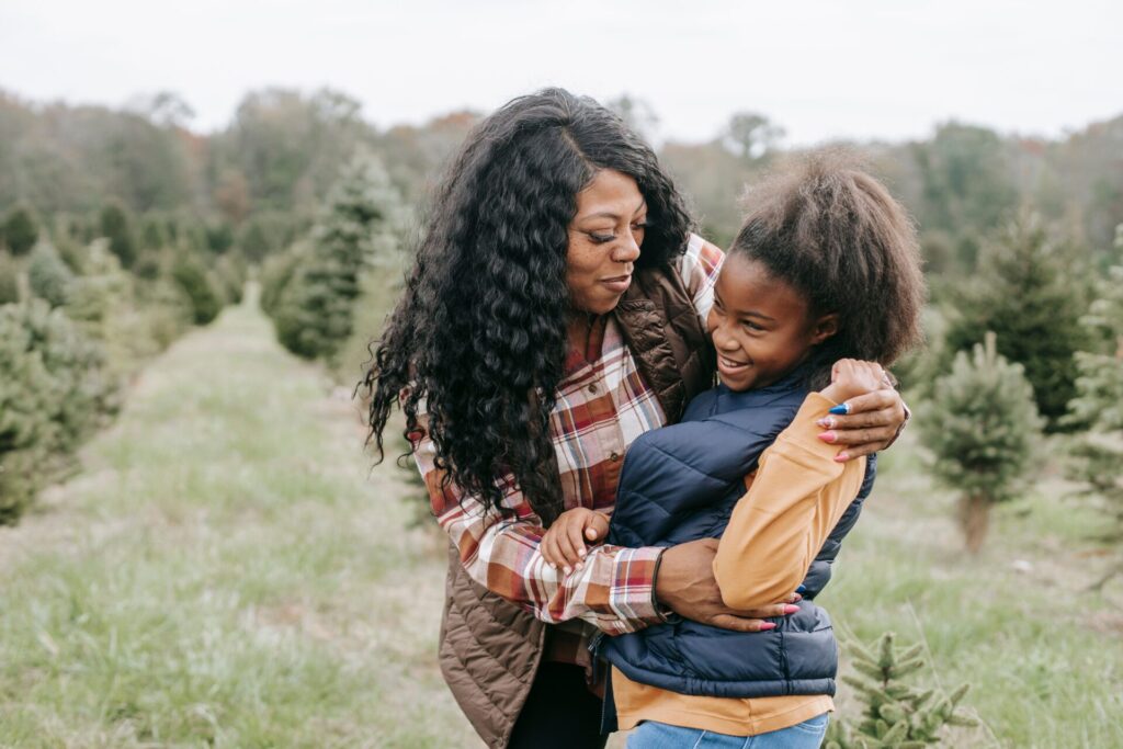 Cheerful African American mum cuddling child while standing close among rows of Christmas trees