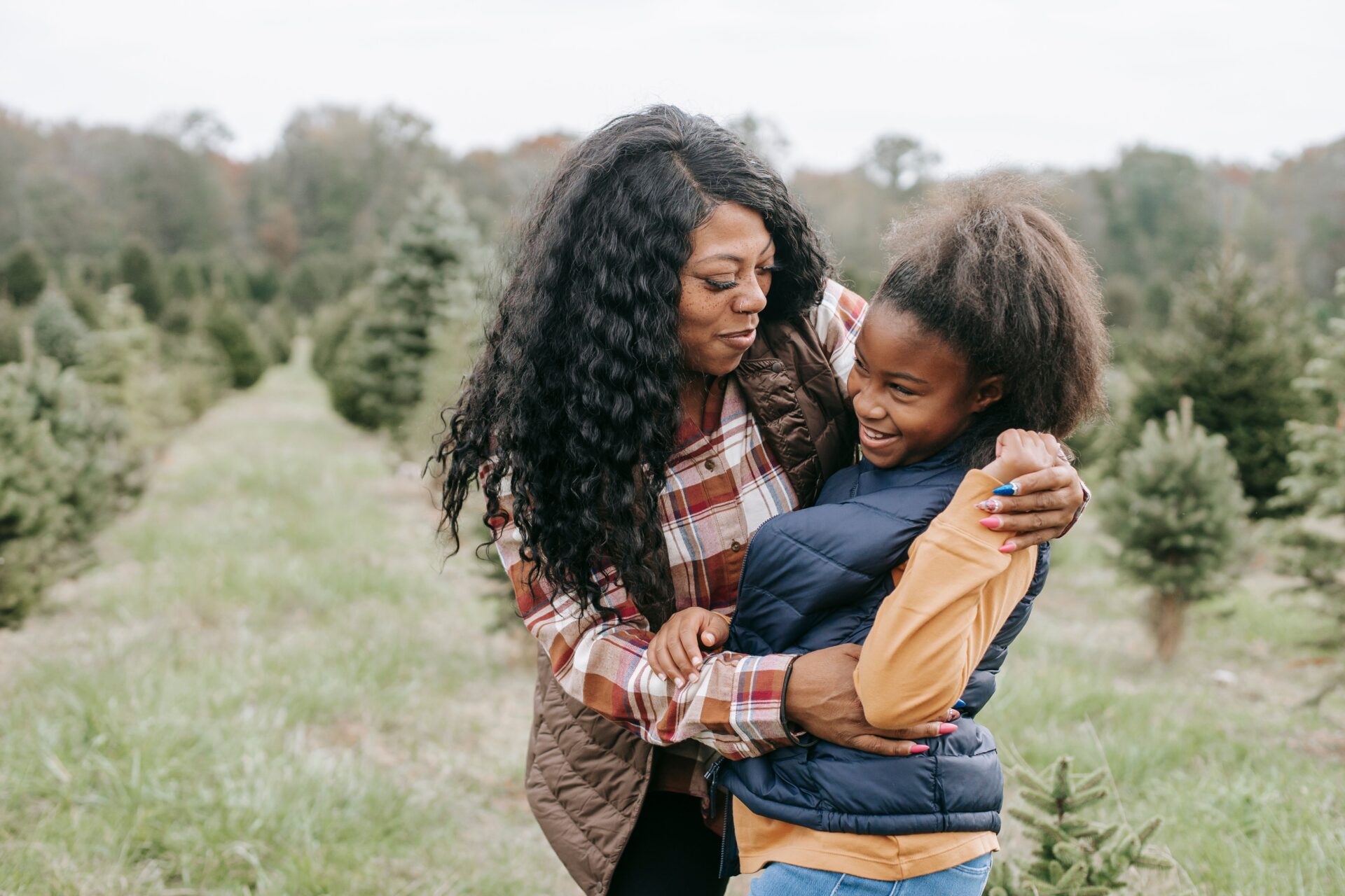 Cheerful African American mum cuddling child while standing close among rows of Christmas trees