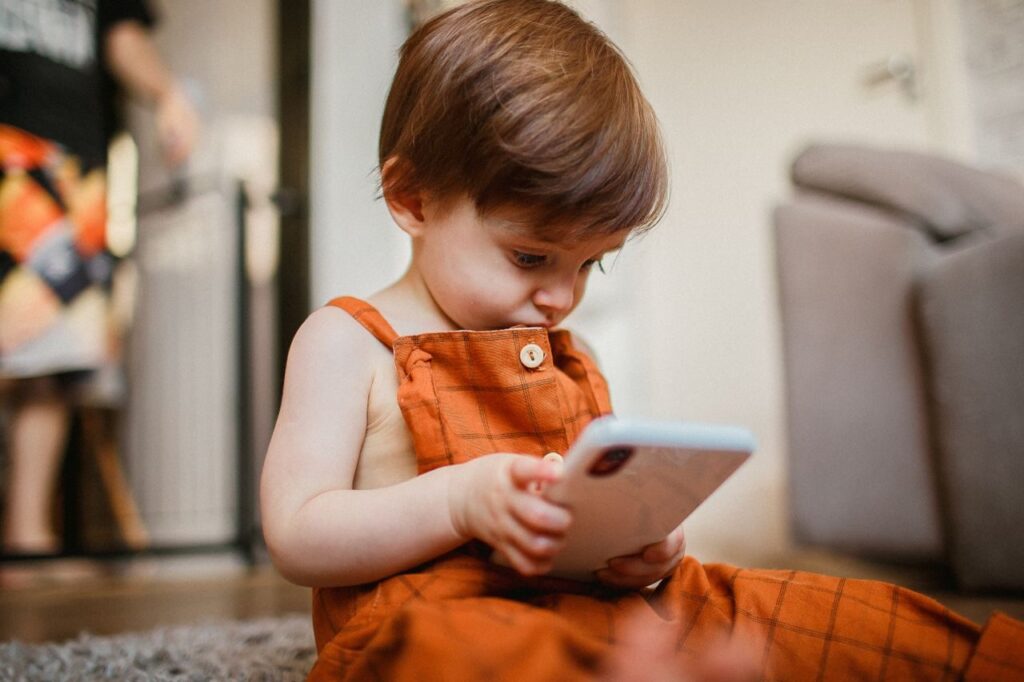 A cute toddler in orange overalls focused on a smartphone indoors, exploring technology playfully.
