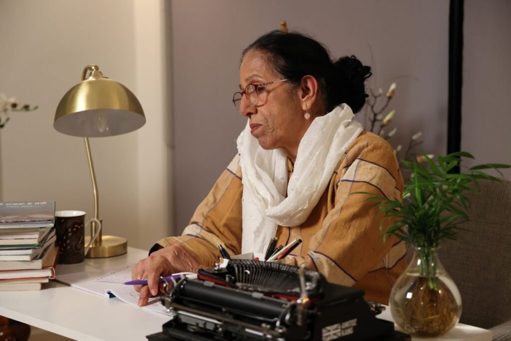 pexels-photo-36687132-36687132 An elderly woman sits at her desk writing with a typewriter in a cozy indoor setting.