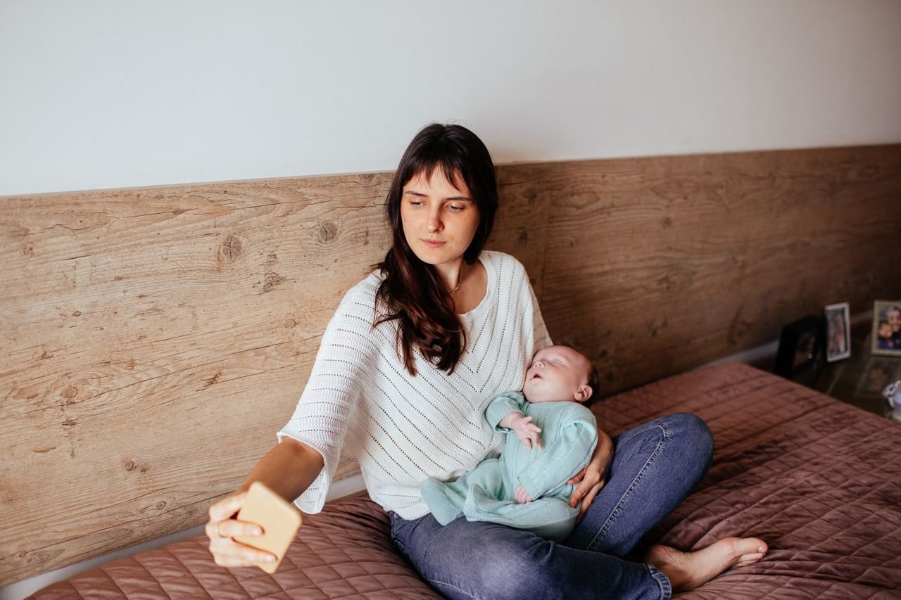 Young mother takes a selfie while holding her sleeping baby in a cozy bedroom setting.