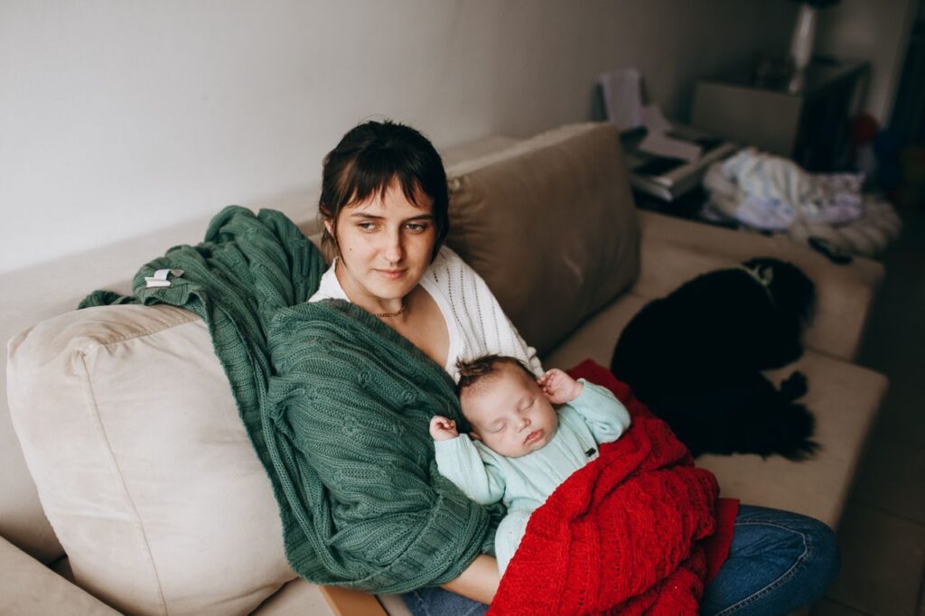 A mother lovingly holds her sleeping baby on a cozy sofa, surrounded by warm blankets.