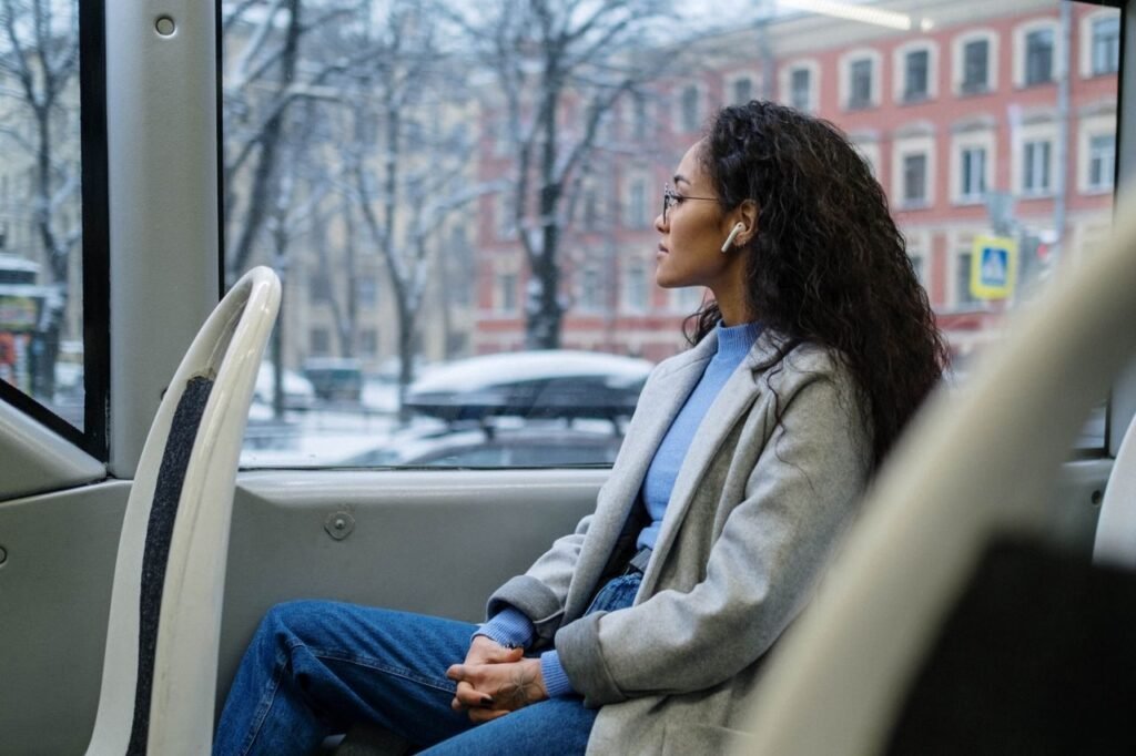 A woman wearing earphones gazes out the window of a bus on a snowy winter day.