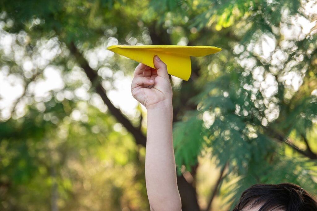 A child playfully raises a yellow paper plane against a lush green background.