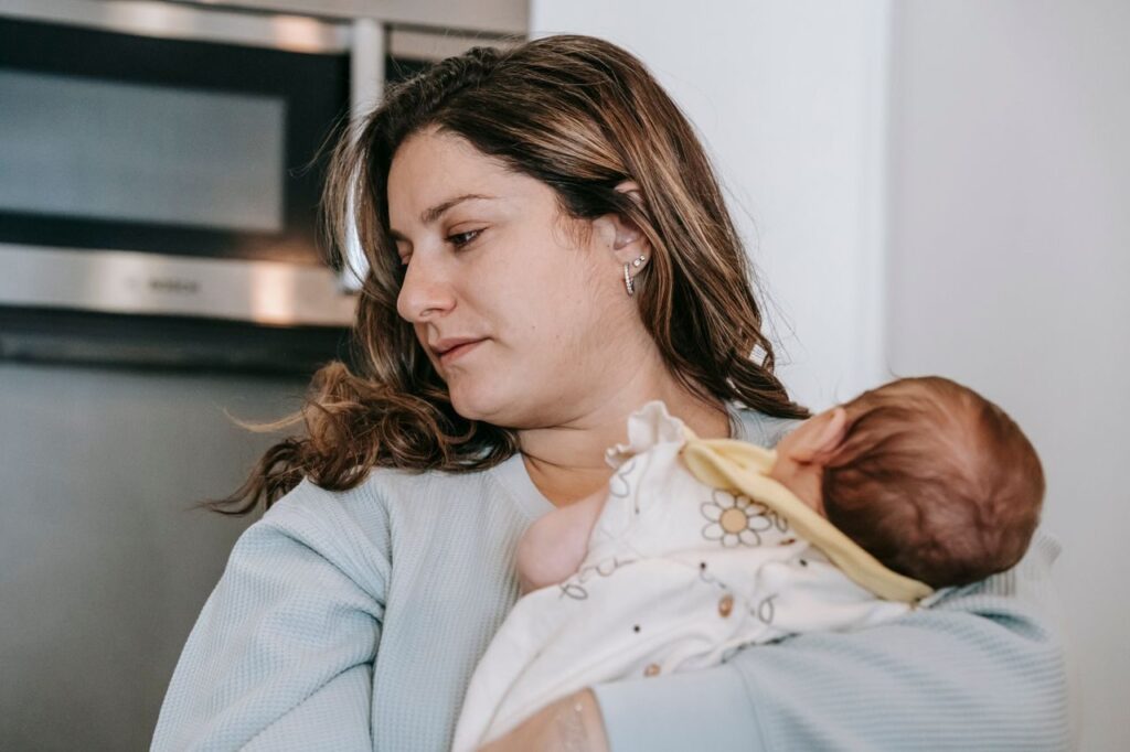 Caring mother with curly hair and cute infant baby in arms standing in light kitchen with microwave oven at home